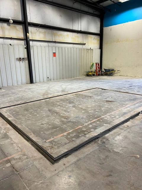 Empty industrial space with a framed outline on the floor, ladder, wheelbarrow, and pale blue walls in the background.