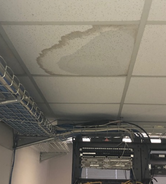 A stained ceiling tile indicates a water leak directly above computer equipment racks in an office setting.